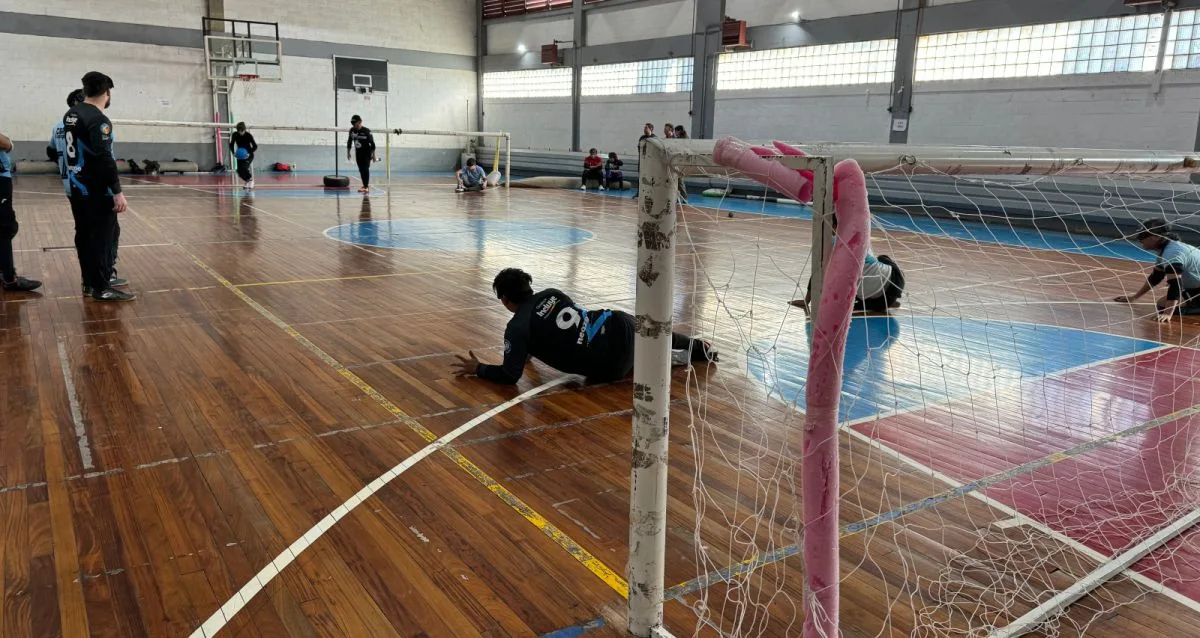 Un grupo de atletas en un gimnasio jugando al goalball. Un jugador se lanza al suelo para bloquear el balón, mientras que otros se encuentran en varias posiciones en la cancha. El gimnasio cuenta con suelos de madera, porterías y ventanales que dejan entrar luz natural.